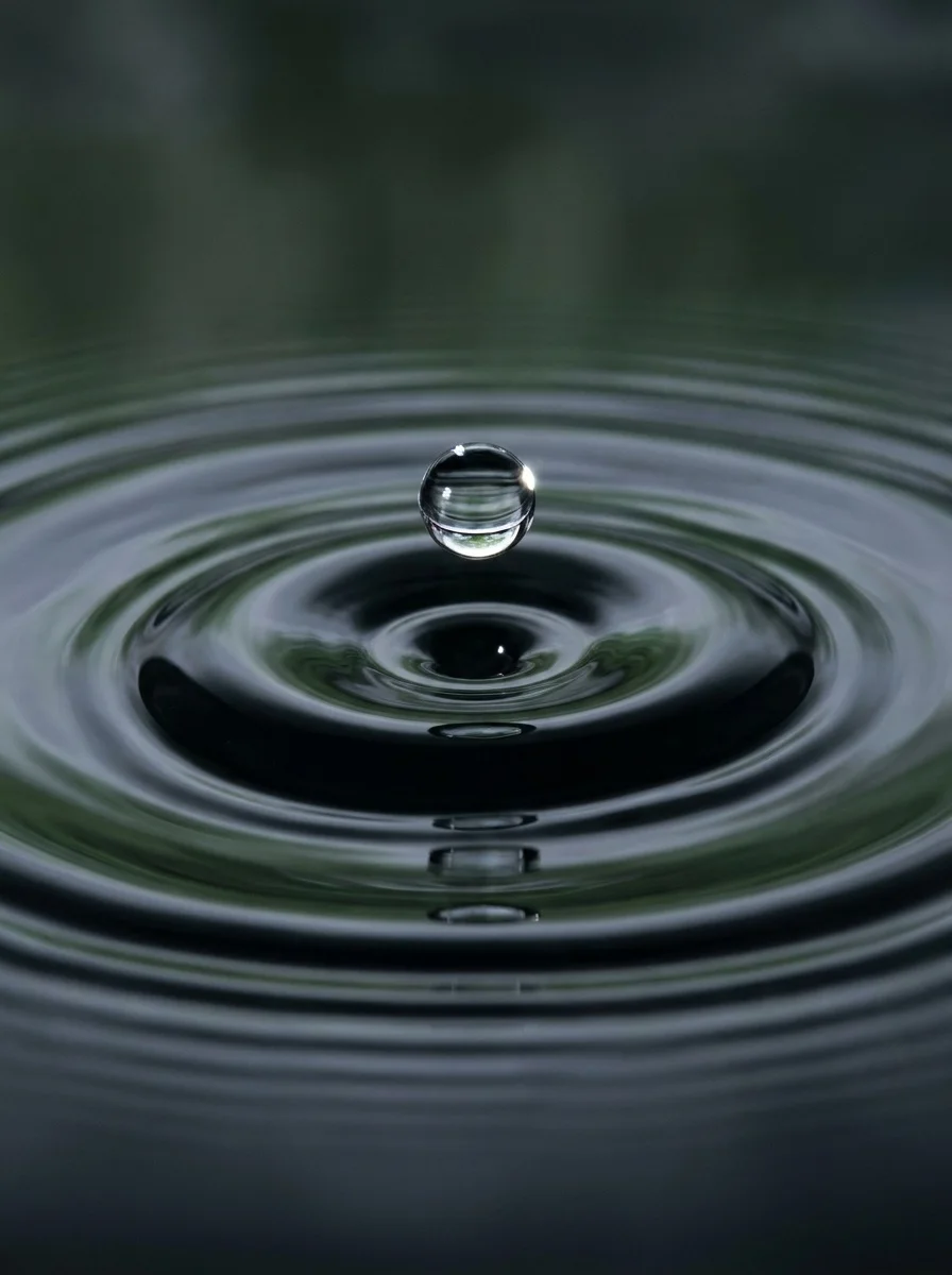 A single clear water droplet meeting a still, dark water surface, with concentric ripples spreading out from it.